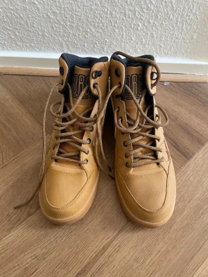 Nike bruna höga kängor - Snygga höga kängor från Nike i ljusbrunt med svarta detaljer och klassisk logga på hälen. Skorna har robust snörning, vadderad insida och slitstark ovandel i syntet och textil. Perfekta för dig som vill ha streetstyle med sportig känsla.