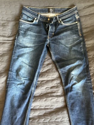 Blå slim fit jeans med slitningar - Snygga blå slim fit jeans med lätt slitna detaljer och klassiska fem fickor. Jeansen har ljusare partier på låren och knäna för en trendig look. Tillverkade i denim med kontrastsömmar och knappgylf.