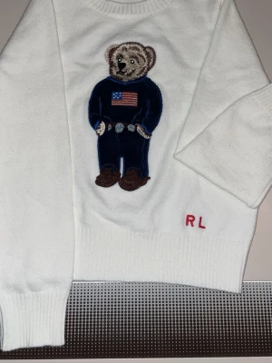 Xs. Vit stickad från Ralph Laruen Björn. - Köpte den som gåva till min lille bror han använde den aldrig säljer pågrund av detta anledning Vit stickad tröja från Ralph Lauren med en ikonisk björn i blå outfit och amerikansk flagga broderad på bröstet. RL-broderi i rött vid nederkanten. Tröjan har rund halsringning och ribbade muddar. Perfekt för dig som gillar klassiska och lekfulla detaljer.