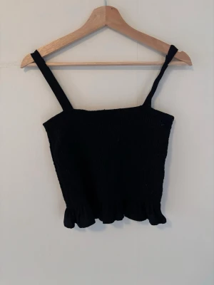 Svart smockad linnetopp H&M - Svart smockad linnetopp från H&M med breda axelband och volangkant nedtill. Toppen är croppad och har en enkel, stilren look som passar perfekt till jeans eller kjol. Materialet är mjukt och stretchigt för en bekväm passform.