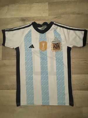Argentina Messi matchtröja Adidas - Säljer en klassisk Argentina matchtröja från Adidas med ljusblå och vita ränder, svarta detaljer och officiella AFA- och FIFA-märken på bröstet. På ryggen står det Messi och nummer 10. Perfekt för fotbollsfans!
