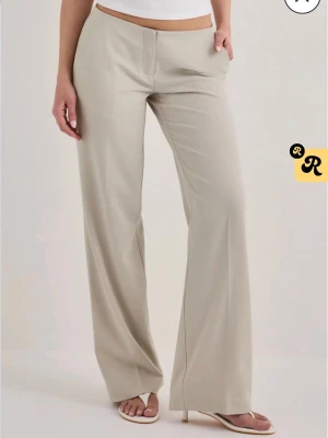 Vida beige kostymbyxa - Snygga vida kostymbyxor i beige med hög midja och pressveck. Byxorna har raka ben och diskreta fickor bak. Perfekta för en clean och stilren look. Tillverkade i ett mjukt, vävt material som ger en skön känsla.