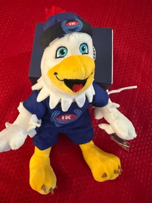 IK Oskarshamn maskot mjukisdjur - Mjukisdjur i form av en glad fågelmaskot klädd i blå IK Oskarshamn-tröja och shorts, med gul näbb och fötter samt blå keps med klubbens logga. Tillverkad i mjukt plyschmaterial och har broderade detaljer som ögon och klubbmärke.