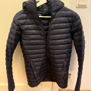 Mörkblå dunjacka Peak Performance 170 - Säljer en mörkblå dunjacka från Peak Performance i storlek 170. Jackan är quiltad med horisontella sömmar, har huva och dragkedja framtill. Obs! Har ett litet litet hål på vänstra armen! Perfekt för kyliga dagar och har en clean, sportig look.