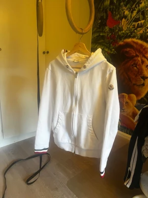 Vit hoodie med dragkedja från Moncler - Säljer en vit hoodie från Moncler med dragkedja framtill och två fickor. Tricolore-detaljer på muddar och huva samt Moncler-logga på ärmen. Klassisk passform och mjukt material, perfekt för en clean och stilren look.