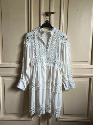 Vit spetsklänning med lång ärm - Superfin vit kortklänning med långa ärmar och detaljerad spets över axlar, bröst och nederkant. Klänningen har en hög krage och v-ringning framtill. Perfekt för sommarens alla tillfällen och ger en romantisk vibe.