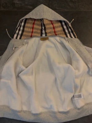 Grå hoodie från Burberry med dragkedja - Snygg grå hoodie från Burberry med dragkedja och klassiskt rutigt foder i huvan. Hoodien har fickor framtill, ribbade muddar och justerbar huva med snören. Tillverkad i mjuk bomullsblandning och har diskret Burberry-logga på bröstet.