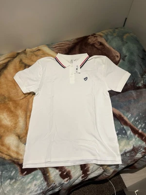 Vit pikétröja med rödblå krage - Snygg vit pikétröja från Oceans Apart med klassisk krage och knappar framtill. Kragen har röda och marinblå ränder för en sportig look. Liten broderad logga på bröstet. Perfekt till jeans eller shorts för en clean stil. Storlek 170. Passar en 14/15 åring och till och med mig som är 180cm lång