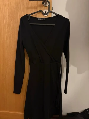 Svart omlottklänning med långa ärmar - Snygg svart omlottklänning från Pieces med långa ärmar och knyt i midjan. Klänningen har en v-ringad hals och är i en kort modell som passar perfekt till många olika tillfällen. Enkel och stilren design som är lätt att matcha.