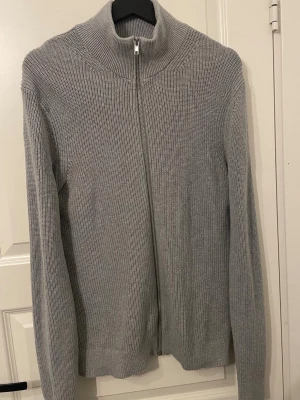 Ljusgrå stickad ziptröja East West - Snygg ljusgrå stickad tröja från EAST WEST med dragkedja framtill och hög krage. Tröjan har lång ärm och ribbstickade detaljer vid ärmslut och nederkant. Perfekt för lager på lager och enkel att matcha med olika outfits.