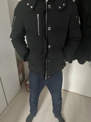 Svart pufferjacka från Moose Knuckles - Snygg svart pufferjacka från Moose Knuckles med stor huva och silverfärgade dragkedjor. Jackan har två framfickor, en ärmficka med metallogo och detaljer i metall. Perfekt för kalla dagar och riktigt street.