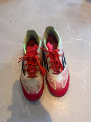 Adidas röda och vita fotbollsskor f50 - Säljer ett par snygga Adidas fotbollsskor i rött och vitt med svarta detaljer och röda skosnören. Skorna har plastdobbar i gult och rött, perfekt för spel på gräsplan. Lätta och smidiga med klassisk Adidas-design och bra grepp.