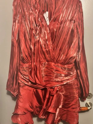 Röd glansig klänning Gina Tricot oanvänd  - Snygg röd klänning i glansigt satinliknande material med långa ärmar och markerad midja. Klänningen har volangdetaljer nertill och en dekorativ knytband vid halsen. Perfekt för dig som vill sticka ut med en trendig och elegant look. Nypris 600 säljer för 400. 
