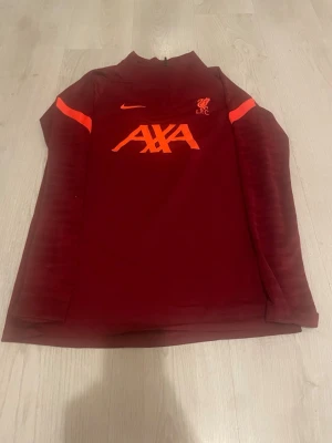 Liverpool träningsjacka Nike röd M - Snygg vinröd träningsjacka från Nike med Liverpool FC-logga och AXA sponsortryck i orange. Tillverkad i Dri-FIT-material som andas, perfekt för träning. Jackan har halv dragkedja och orange detaljer på axlarna.