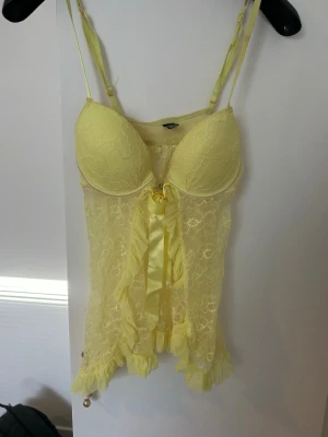 Gul spets med rosett - Söt gul topp i spets med vadderade kupor och justerbara axelband. Dekorerad med en stor satinrosett framtill och volangkant nedtill. Tillverkad i 88% nylon och 12% elastan för stretchig och bekväm passform.