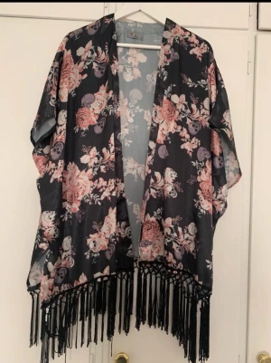 Bohoo kimono med fransar - Säljer en svart kimono med blommor och dödskallemotiv . Toppen är öppen framtill, har korta ärmar och långa svarta fransar längs nederkanten. Perfekt för att lyfta vilken outfit som helst med en bohemisk touch. Storlek xs-s, passar även m . Fint skick ! 