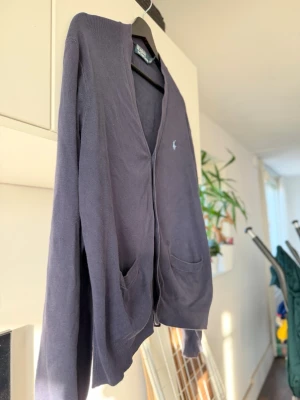 Mörkblå cardigan från Polo Ralph Lauren - Klassisk mörkblå cardigan från Polo Ralph Lauren med V-ringning och knäppning framtill. Tröjan har två fickor och den ikoniska broderade loggan på bröstet. Perfekt för lager på lager och enkel att matcha med olika outfits. Knappen är sönder till viss del, men fungerar ändå.