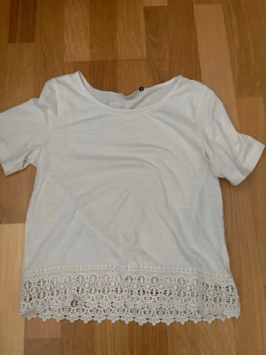 Vit topp med spetskant - Superfin vit topp med korta ärmar och rund halsringning. Nedtill har toppen en bred, detaljerad spetskant som ger en söt och romantisk känsla. Perfekt att styla med jeans eller kjol för en fräsch look.