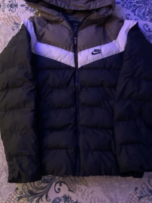 Svart pufferjacka från Nike med huva - Snygg svart pufferjacka från Nike med vita och grå detaljer över bröstet och axlarna. Jackan har dragkedja framtill, huva och klassisk Nike-logga på vänster sida. Perfekt för kalla dagar när du vill hålla stilen.