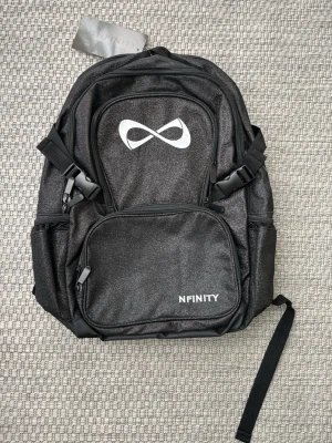 Svart ryggsäck från Nfinity - Snygg svart ryggsäck från Nfinity med vit logga framtill. Ryggsäcken har flera fack med dragkedja, justerbara axelband och sidofickor. Perfekt för träning eller sportaktiviteter. Tillverkad i slitstarkt syntetmaterial. Helt ny men var trasig när jag fick den,(se bild 3). Tror det går att fixa enkelt. Den kostade 2000, med frakt osv.