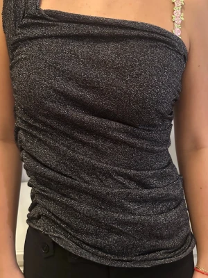 Glittrig svart oneshoulder topp - Säljer en glittrig svart oneshoulder topp. Toppen har en tight passform och är perfekt för dig som vill sticka ut. Materialet är stretchigt och skimrar snyggt i ljuset. Från Ann-Sofie Back