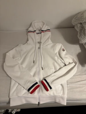 Moncler kofta  - Snygg vit hoodie från Moncler med hel dragkedja framtill och huva med snörning. Ribbade muddar och huva har röd, vit och marinblå rand. Moncler-logga på ärmen. Perfekt för en clean och sportig look.