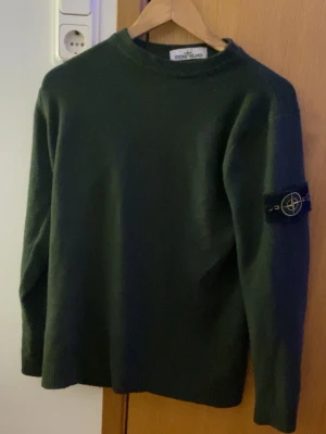 Mörkgrön stickad tröja Stone Island - Snygg mörkgrön stickad tröja från Stone Island med klassisk patch på vänster ärm. Tröjan har rund halsringning och ribbade muddar vid ärmslut och nederkant. Perfekt för dig som gillar streetwear och vill ha något stilrent.
