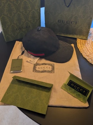 Svart Gucci keps  - Säljer en svart keps från Gucci med klassiskt GG-monogram i tyget och justerbar rem bak. Kepsen har en röd och grön rand på sidan, vilket är Guccis signaturfärger. Materialet är canvas med läderdetaljer. Perfekt för dig som vill ha en exklusiv och trendig accessoar.