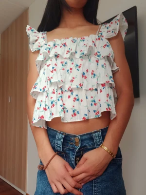 Vit blommig volangtopp med axelband - Supersöt vit topp med blommigt mönster i rött och blått. Toppen har volanger i flera lager och smala axelband med volangdetaljer. Croppad modell som passar perfekt till höga jeans eller kjol. Lätt och luftig för varma dagar.