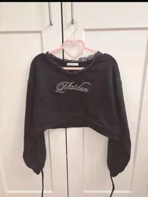 Svart croppad topp med spets och strass - Svart croppad topp från DMLMillor med lång ärm och spetsdetalj vid halsen. Texten 'Maiden' i strass framtill ger en extra touch. 