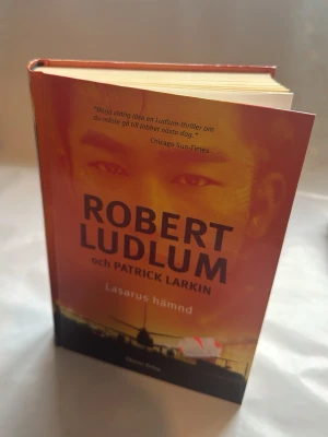 Lazarus hämnd av Ludlum & Larkin - En spännande skönlitterär bok med hårda pärmar, skriven av Robert Ludlum och Patrick Larkin. Omslaget är rött och svart med ett flygplan på ryggen. Perfekt för dig som gillar action och thrillers.