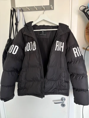 Svart pufferjacka från Hoodrich - Svart pufferjacka från Hoodrich med text på framsidan, armarna och baksidan. Jackan har en stor huva, dragkedja framtill och två sidofickor. Perfekt för kalla vinterdagar och ger en cool streetwear-look.