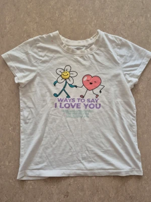 T-shirt - Vit t-shirt i bomull med korta ärmar och ett gulligt tryck framtill av en blomma och ett hjärta som håller hand. Under motivet står texten 'Ways to say I love you' i lila. Perfekt för dig som gillar söta och lekfulla plagg. Finns rosa fläck.
