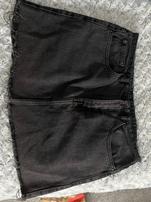 Svart jeanskjol med rå kant - Säljer en svart jeanskjol med rå, fransig kant nertill. Kjolen har klassisk femficksdesign och bälteshällor i midjan. Perfekt för dig som gillar en enkel men edgy stil.