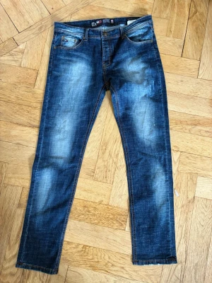Blå slitna jeans med raka ben - Säljer ett par blå jeans med snyggt sliten tvätt och raka ben. Jeansen har klassiska fem fickor, kontrastsömmar och en cool faded effekt framtill och baktill. Perfekta för en avslappnad och trendig look.