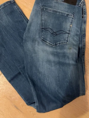 Blå slim fit jeans Replay Anbass - Snygga blå slim fit jeans med ljus tvätt och broderad bakficka. Jeansen har klassisk femficksdesign och smal passform som sitter tajt längs benen. Perfekta för en avslappnad och trendig look.