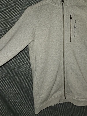 Ljusgrå hoodie från Sail Racing - Ljusgrå hoodie från Sail Racing med hel dragkedja framtill och en bröstficka med dragkedja. Diskret logga på bröstet och klassisk huva. Tillverkad i mjukt sweatshirtmaterial, perfekt för en avslappnad stil. Är öppen för prisförslag vid snabb försäljning.