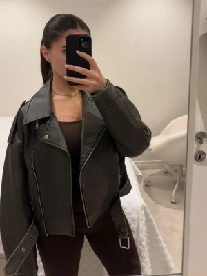 Svart oversized skinnjacka - Säljer en svart oversized skinnjacka med breda slag, dragkedja och bälte i midjan. Jackan har coola detaljer som axelklaffar och silverfärgade spännen. Perfekt för dig som vill ha en trendig och edgy look.