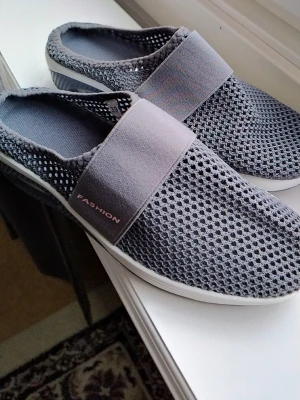 Grå mesh-tofflor med bred resår - Säljer ett par gråa tofflor från Fashion med luftig mesh-ovandel och bred resår över foten. De har vit sula och öppen häl, perfekt för dig som vill ha något lätt och bekvämt att glida i. Stilen är sportig och modern med diskret logga på sidan.helt nya 