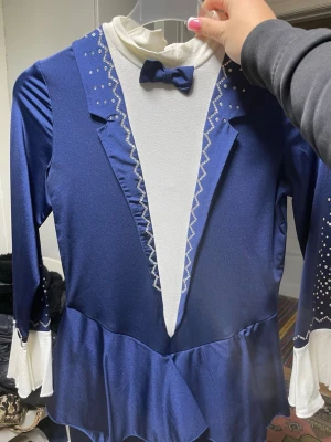 Blå och vit maskeradkostym med paljetter - Snygg maskeradkostym i blått och vitt med lång ärm, paljettdekor och fastsydd fluga. Kostymen har vita manschetter och detaljerade mönster längs kragen och ärmarna. Perfekt för cosplay eller temafest! Användes tidigare som konståknings dreckt, använd 5-7 gånger 💕(hand gjord) storlek M/S 