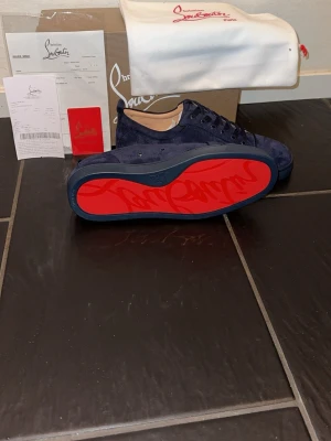 Mörkblå sneakers från Christian Louboutin - Snygga mörkblå sneakers i mocka från Christian Louboutin med den ikoniska röda sulan och diskret logga. Skorna har lågt skaft, matchande mörkblå snörning och beige innersula. Perfekta för dig som vill sticka ut med exklusiv stil.