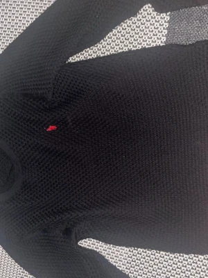 Svart stickad tröja Polo Ralph Lauren - Klassisk svart stickad tröja från Polo Ralph Lauren med rund halsringning och röd broderad logga på bröstet. Tröjan har en grovstickad struktur och ribbade muddar vid hals, ärmslut och nederkant. Perfekt för dig som gillar stilrena plagg.