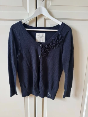 Mörkblå kofta Abercrombie & Fitch - Mörkblå kofta med dekorativa blommor vid ena axeln från Abercrombie & Fitch. Koftan har v-ringning, knäppning framtill och långa ärmar. 