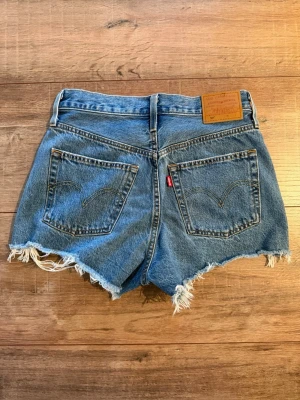 Levi's 501 blå jeansshorts med fransar  - Säljer ett par klassiska blå jeansshorts från Levi's med hög midja och råa, fransiga kanter. Shortsen har slitna detaljer framtill, fem fickor och Levi's ikoniska läderpatch bak. Perfekta för sommaren och ger en avslappnad vibe.