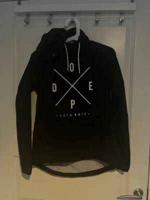 Svart vindjacka från Dope - Svart vindjacka från Dope med stor vit logga och tryck framtill. Jackan har huva med dragsko, dragkedjor i sidorna och en stor magficka. Oanvänd eftersom att den var för liten för mig och jag köpte en annan! Har inget kvitto kvar därav priset!!☺️