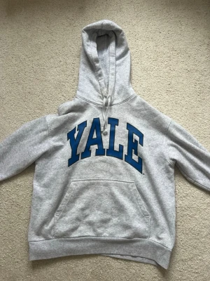 Grå Yale hoodie med blå text - Snygg grå hoodie med stor blå YALE-text på bröstet. Klassisk känguruficka framtill och justerbar huva med dragsko. Tillverkad i mjuk bomullsblandning som är skön att ha på sig. Perfekt för dig som gillar college-stil och vill ha en avslappnad look.