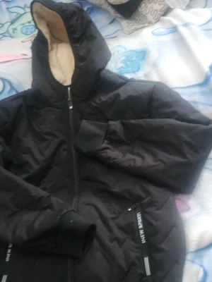 Svart teddyjacka med dragkedja - Mysig svart teddyjacka från New Sports med mjukt beige foder. Jackan har dragkedja framtill, ribbade muddar och två fickor. Perfekt för kalla dagar när du vill hålla dig varm och bekväm.