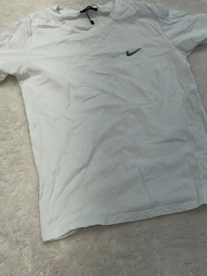 Vit Nike t-shirt med logga - Klassisk vit t-shirt från Nike med liten svart swoosh-logga på bröstet. T-shirten är kortärmad och har en enkel, stilren design. Perfekt för dig som gillar sportiga och avslappnade plagg. Tillverkad i mjuk bomull för skön känsla.