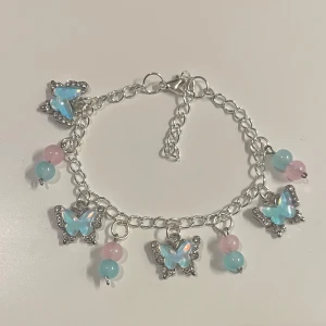Silverfärgat armband med fjärilar - HANDGJORT armband i silver med fem blå skimrande fjärils berlocker och pärlor i pastellrosa och ljusblått. Länkkedja med justerbar längd och karbinlås.