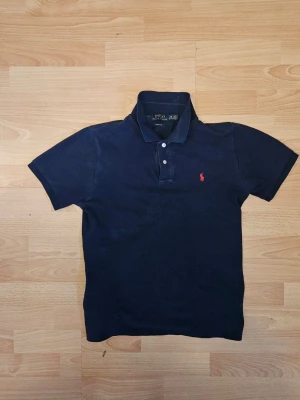 Marinblå pikétröja från Ralph Lauren - Klassisk marinblå pikétröja från Ralph Lauren med korta ärmar och krage. Tröjan har två knappar framtill och den ikoniska röda broderade loggan på bröstet. Perfekt till jeans eller shorts för en clean och stilren look.